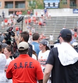 A lively gathering in an outdoor sports venue with a group of people, some wearing matching shirts labeled 'National Champions.' Several individuals are holding cameras and microphones, indicating a media presence. The background shows blurred spectators sitting on bleachers.