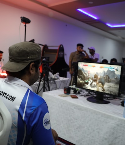 A group of people is gathered in a room watching and participating in a video game competition. Players and spectators are focused on the monitors displaying a fighting game. The setting includes tables with equipment and chairs arranged around the venue, with a noticeable intensity in the participants’ engagement.